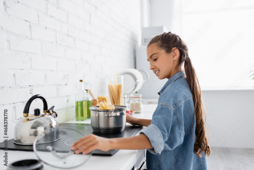 Side view of positive kid taking cap near saucepan with macaroni on stove in kitchen Side view of positive kid taking cap near saucepan with macaroni on stove in kitchen
