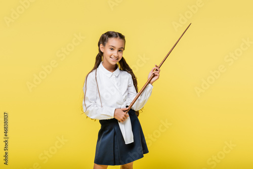 positive schoolgirl with pointer looking at camera isolated on yellow
