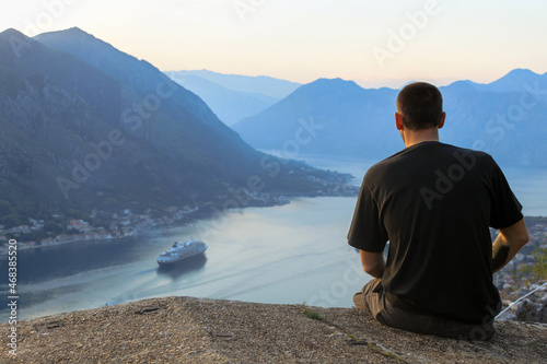 A man in a black T-shirt sits looking down on a cruise ship in a sea bay. Against the backdrop of a mountain. Pre-sunset time. The concept of tourism in Montenegro.