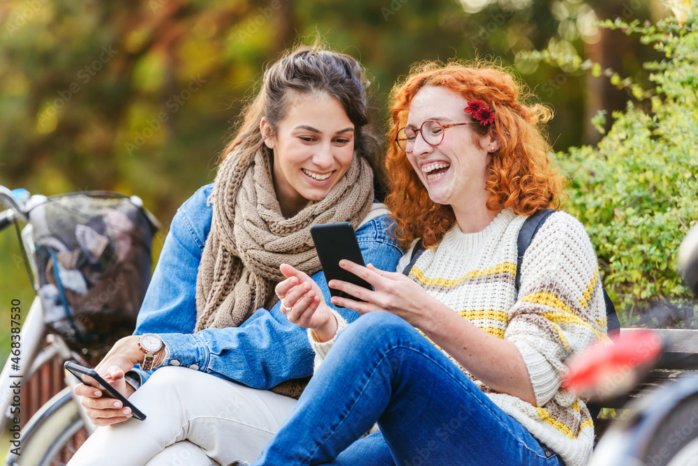 Cheerful female friends sitting on a bench in a park talking, smiling, using smartphones Cheerful female friends sitting on a bench in a park talking, smiling, using smartphones