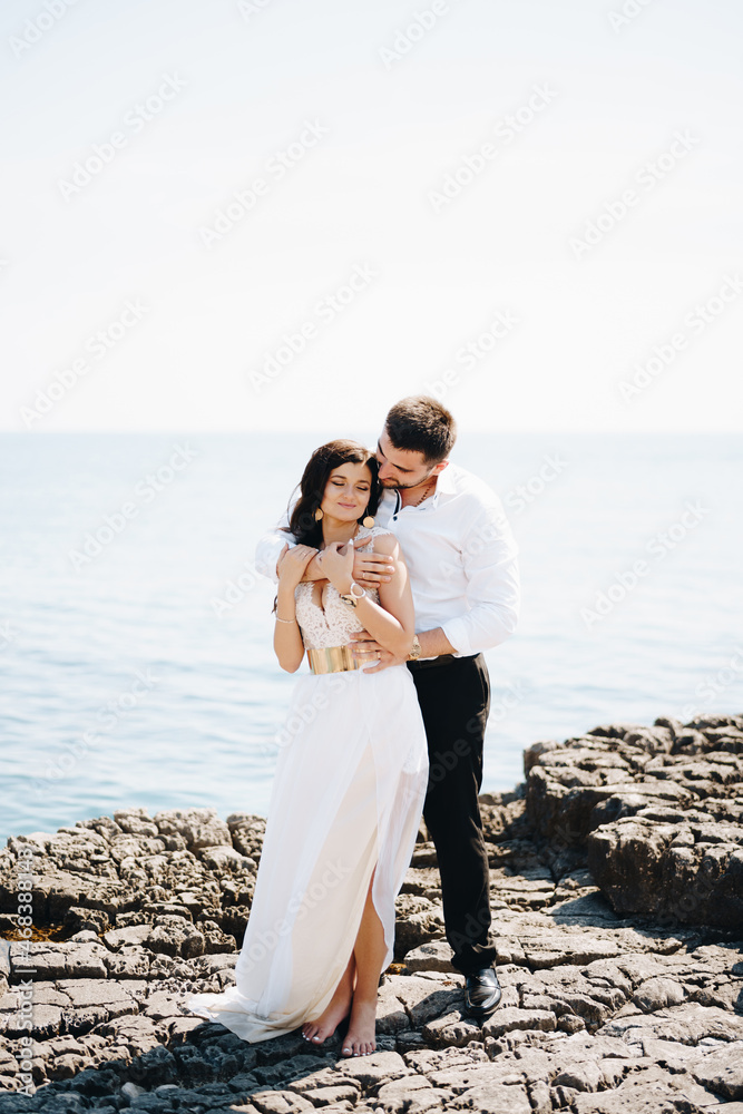 The bride and groom stand hugging on the rocky shore 