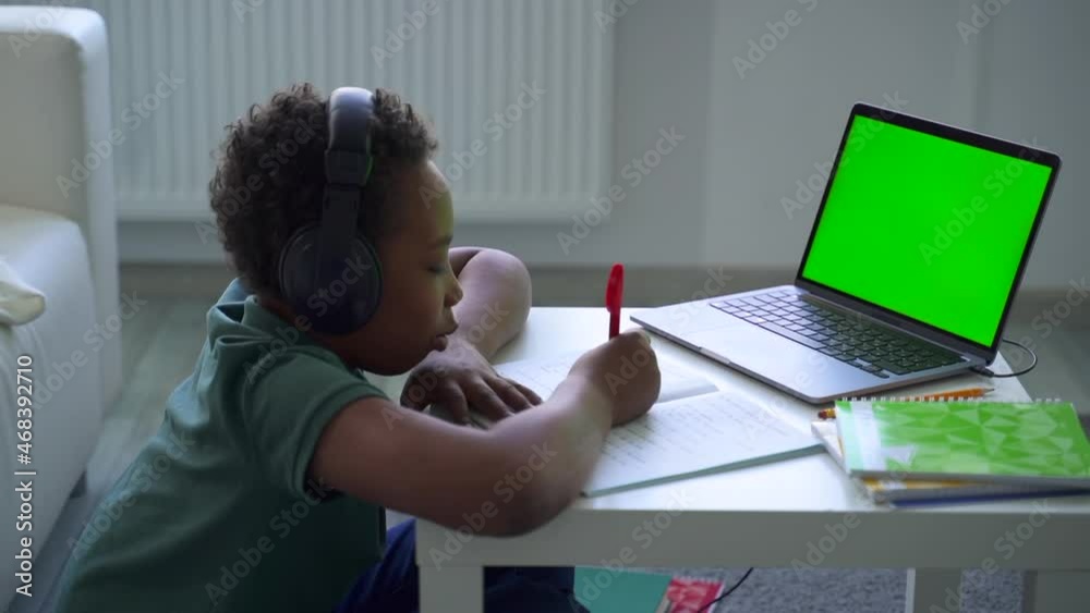 Distant home schooling of junior children. african-american boy kid student with headset writes spbd and listens to teacher at videochat via laptop in room Distant home schooling of junior children. african-american boy kid student with headset writes spbd and listens to teacher at videochat via laptop in room