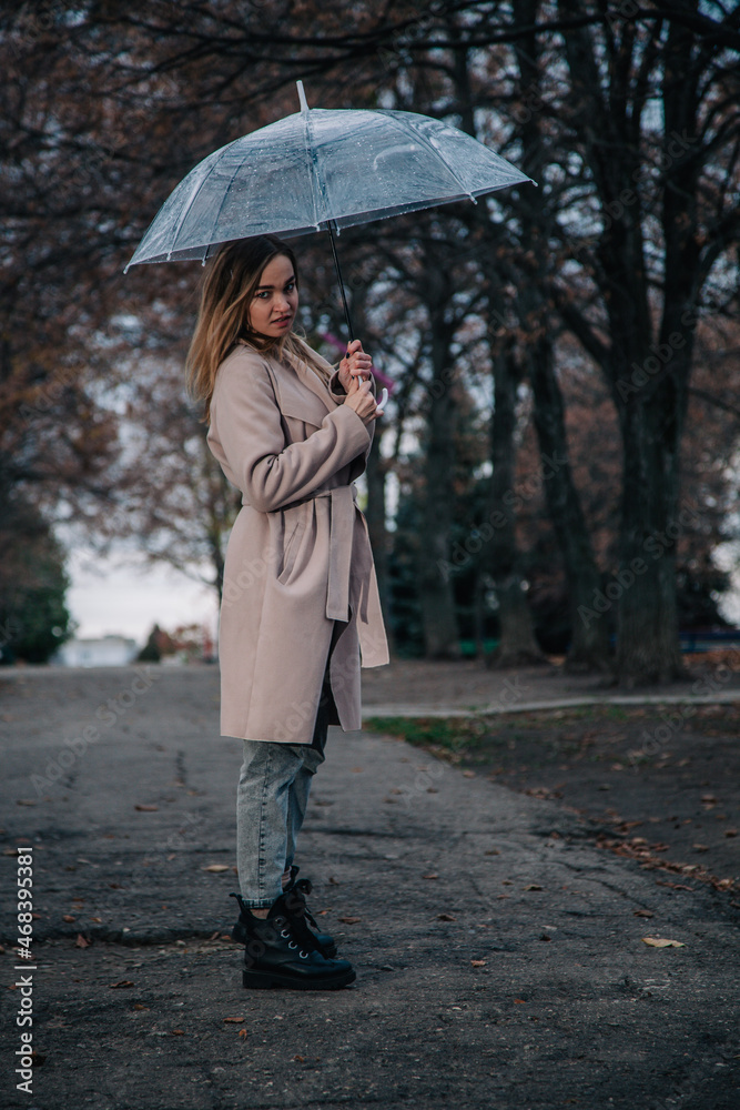 A young, slender, beautiful girl in a coat walks in the rain with a transparent umbrella in the park A young, slender, beautiful girl in a coat walks in the rain with a transparent umbrella in the park