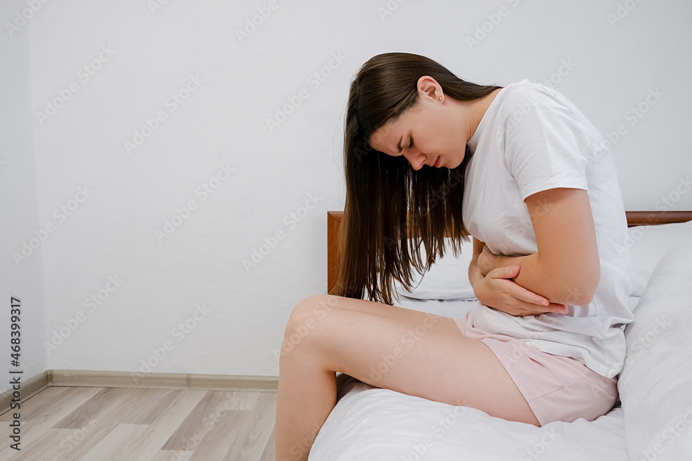 Upset young caucasian woman sitting on white bed at home holding belly suffering from stomach pain feeling painful discomfort hurt abdomen ache having pancreatitis problem. Indigestion symptom concept Upset young caucasian woman sitting on white bed at home holding belly suffering from stomach pain feeling painful discomfort hurt abdomen ache having pancreatitis problem. Indigestion symptom concept