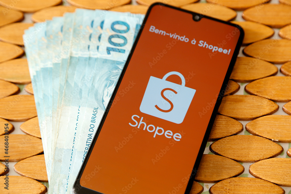 Shopee logo on mobile screen and Brazilian money bills Photos | Adobe Stock
