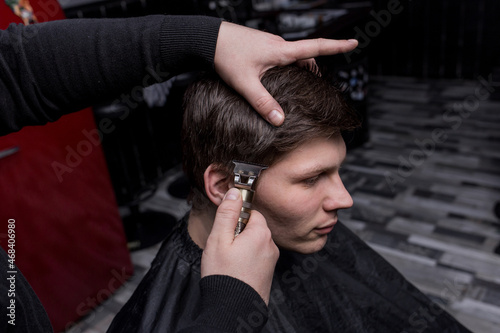 The hand of a barber or hairdresser cuts the dark long hair of the guy's client on his temple. Hairdressing