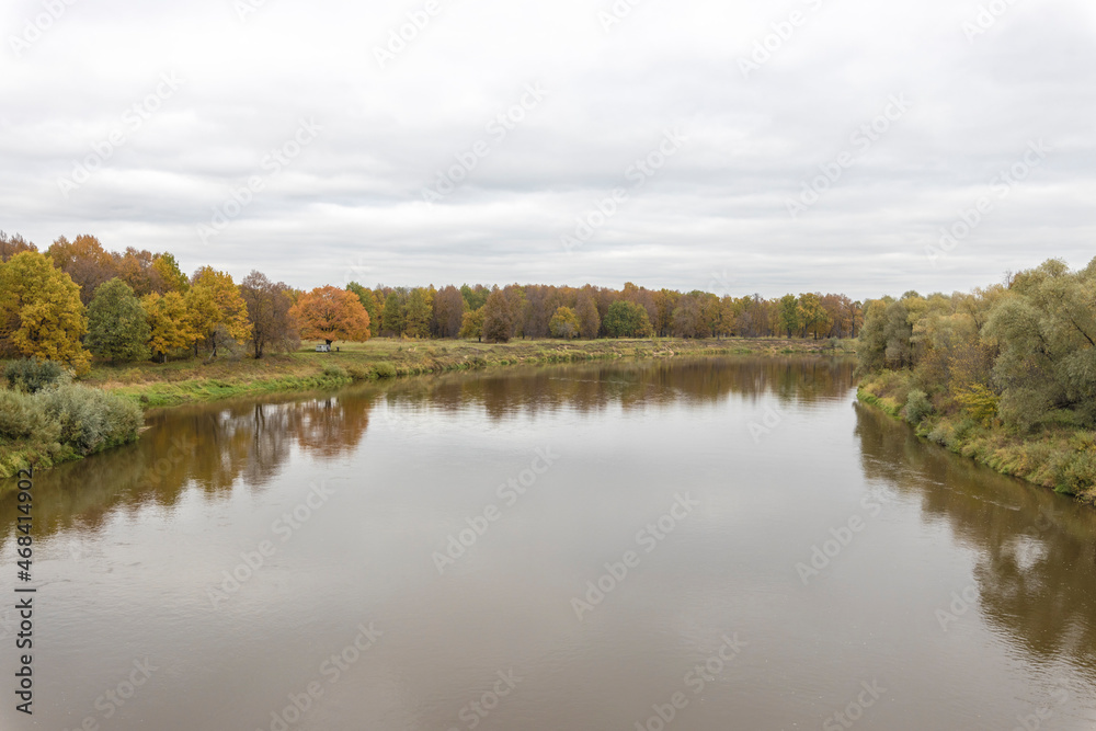 Autumn landscape with a river. Cloudy autumn day by the river. View of the river with trees and bushes on the bank. Bright colors of autumn on a cloudy day.
