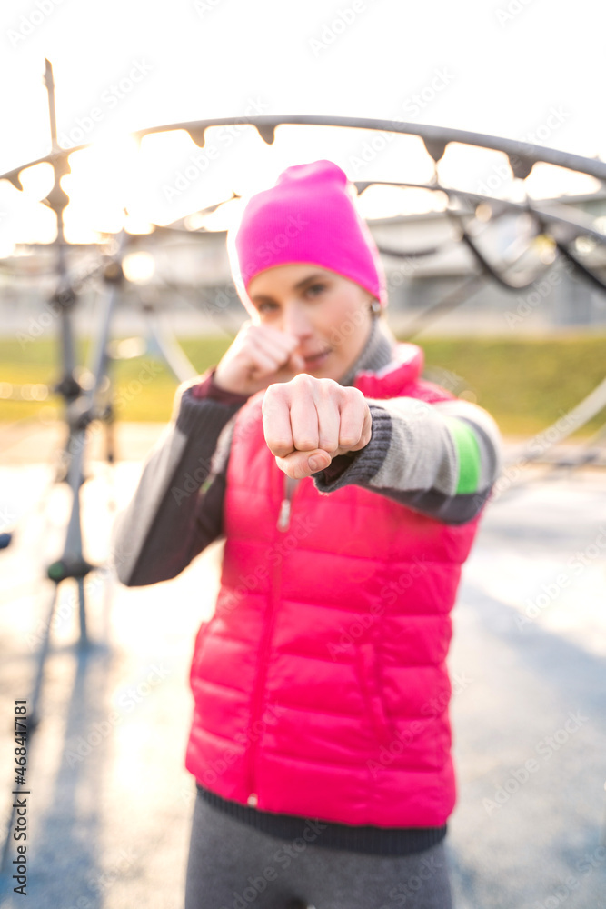 Obraz premium Young caucasian fit woman in pink fitness clothes boxing with hands in fists and makes punches on modern athletic field outdoors on sunrise on cold autumn, winter or spring morning. Sports, training