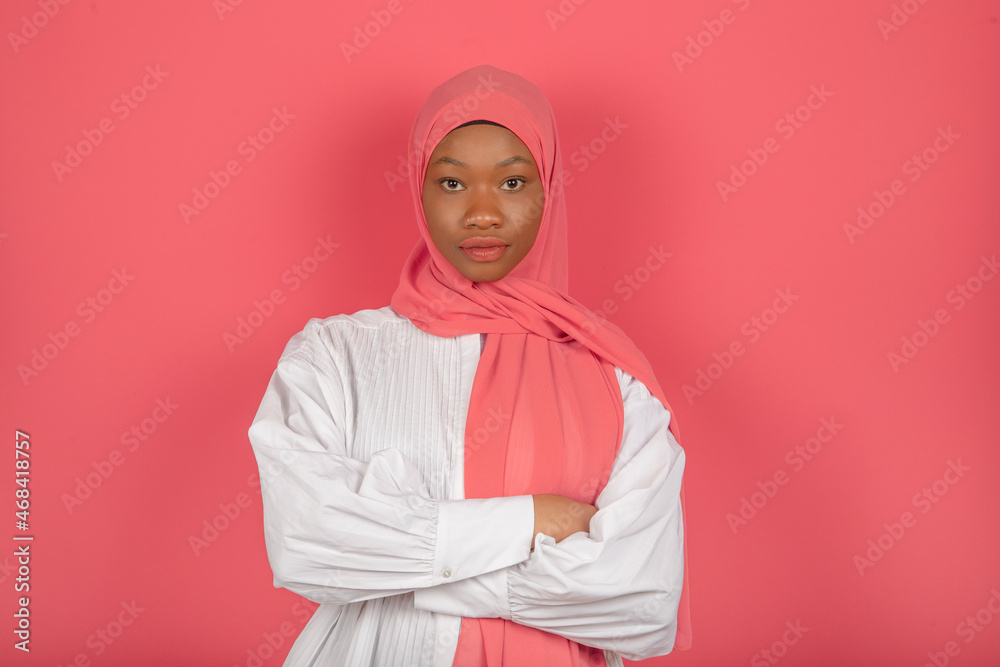 Arab woman with serious and calm expression with dark skin, wrapped in pink veil, wears white shirt on pink studio wall. Arab woman with serious and calm expression with dark skin, wrapped in pink veil, wears white shirt on pink studio wall.