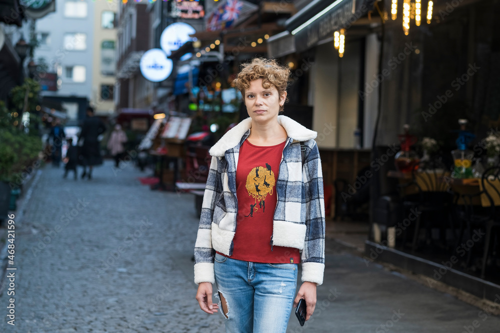 Young fashionably and stylishly dressed woman in a short plaid jacket and jeans with short curly hair is walking along a city street in autumn