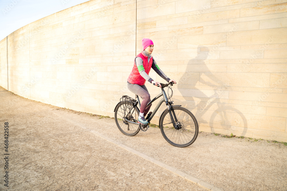 Naklejka premium Young caucasian active woman in warm pink fitness clothes riding bike outdoors near wall on streets or athletic sports ground on winter or spring early morning sunrise. Active lifestyle, transport