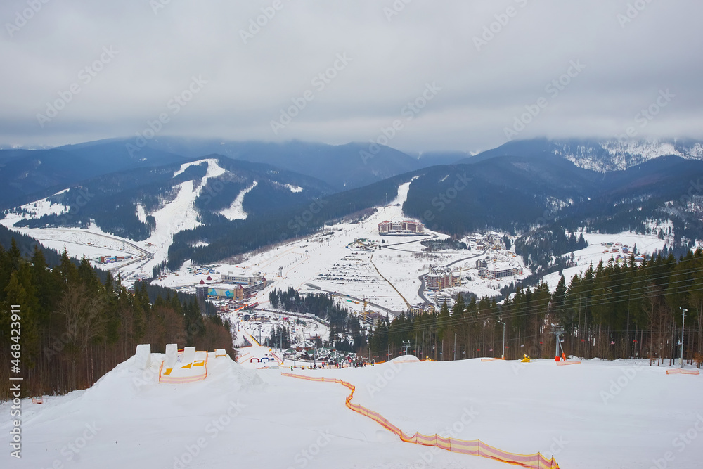 Fototapeta premium Ski resort in beautiful sunset light. View from the top: ski tracks, pine tree forest and mountains in the background.