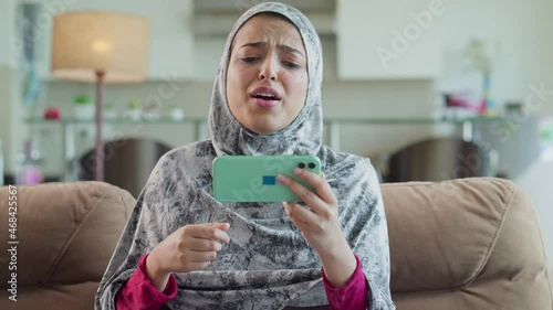 A Modern young beautiful Arabic muslim or middle eastern woman wearing hijab sitting on couch and anxiously watching a game of football or soccer match on a Smart phone in an interior house setup photo