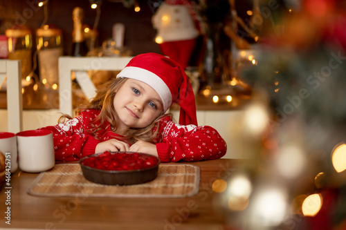 a cute little girl child in a red sweater and a Santa Claus hat is in the kitchen drinking tea with a pie and waiting for the new year or Christmas