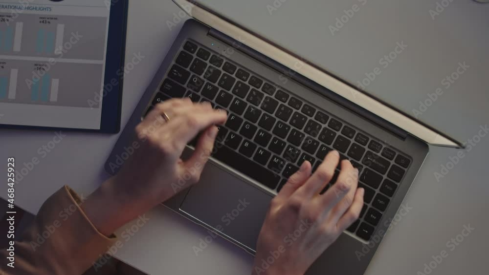 Top-view close-up of unrecognizable female hands typing on laptop keyboard on desk with financial documents