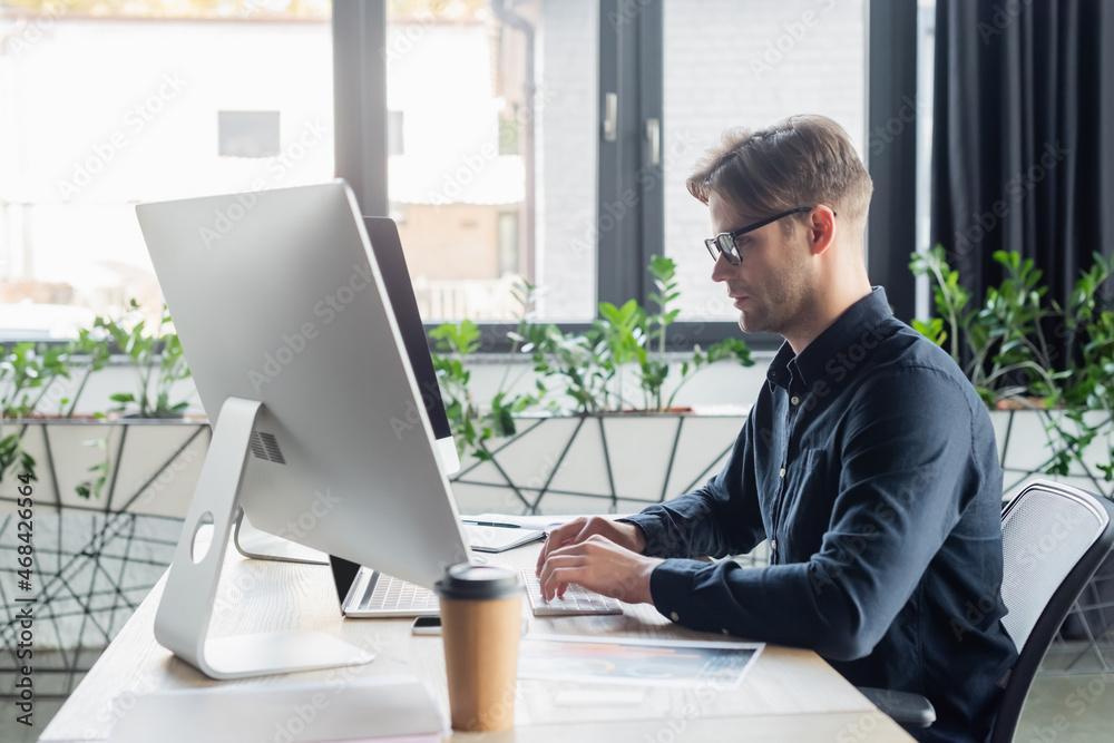 Side view of programmer using computers near blurred document and coffee in office