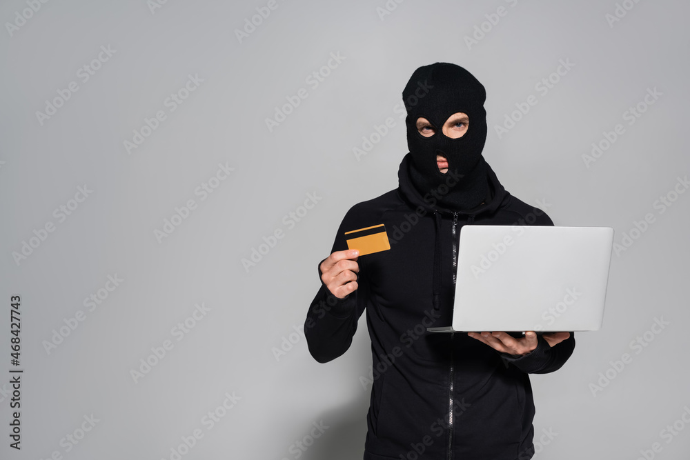 Hacker in balaclava holding credit card and laptop on grey background Hacker in balaclava holding credit card and laptop on grey background