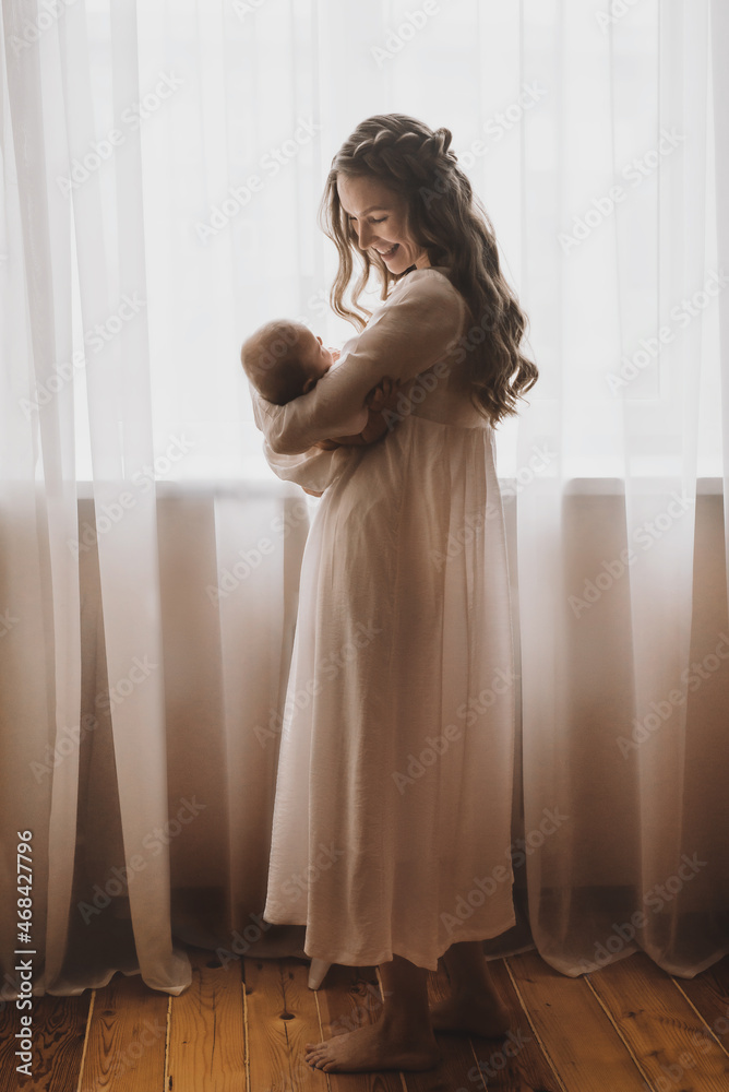 Fototapeta premium Beautiful young mother holds newborn baby girl in white lighter room at home. Tenderness cozy atmosphere