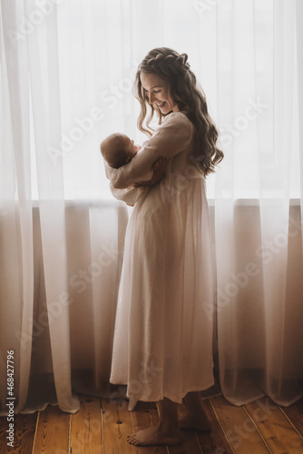 Beautiful young mother holds newborn baby girl in white lighter room at home. Tenderness cozy atmosphere
