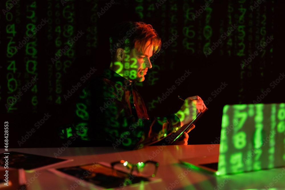 Side view of programmer writing on clipboard near laptop with reflection of computer code on black background