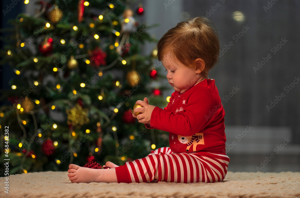 A small child with red hair and a red Christmas costume sits at home against the background of a Christmas tree and plays with Christmas decorations A small child with red hair and a red Christmas costume sits at home against the background of a Christmas tree and plays with Christmas decorations