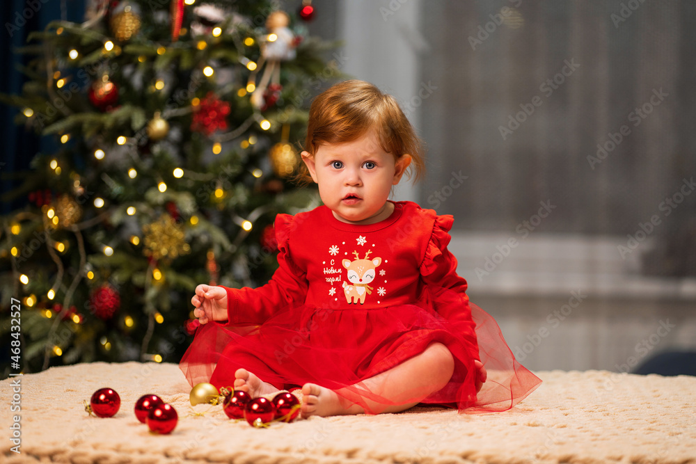 A baby with red hair sits in a New Year's red dress at home against the background of a Christmas tree. Inscription on the dress: Happy New Year! A baby with red hair sits in a New Year's red dress at home against the background of a Christmas tree. Inscription on the dress: Happy New Year!