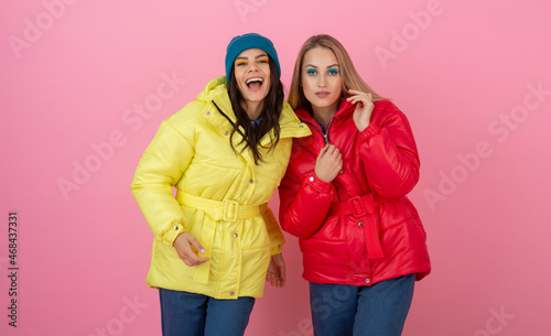 two excited attractive girl friends active women posing on pink background in colorful winter down jacket of bright red and yellow color having fun together, warm coat fashion trend