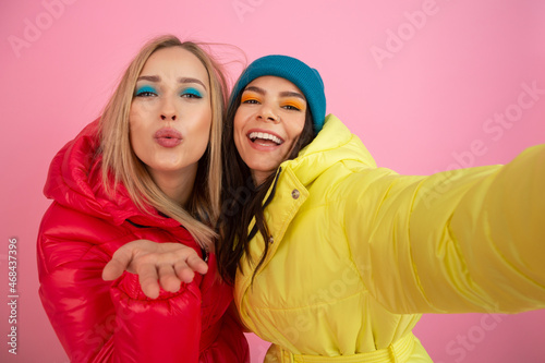 two attractive girl friends women taking selfie photo on pink background in colorful winter jacket of bright red and yellow color having fun together, warm coat sportswear fashion trend, crazy funny