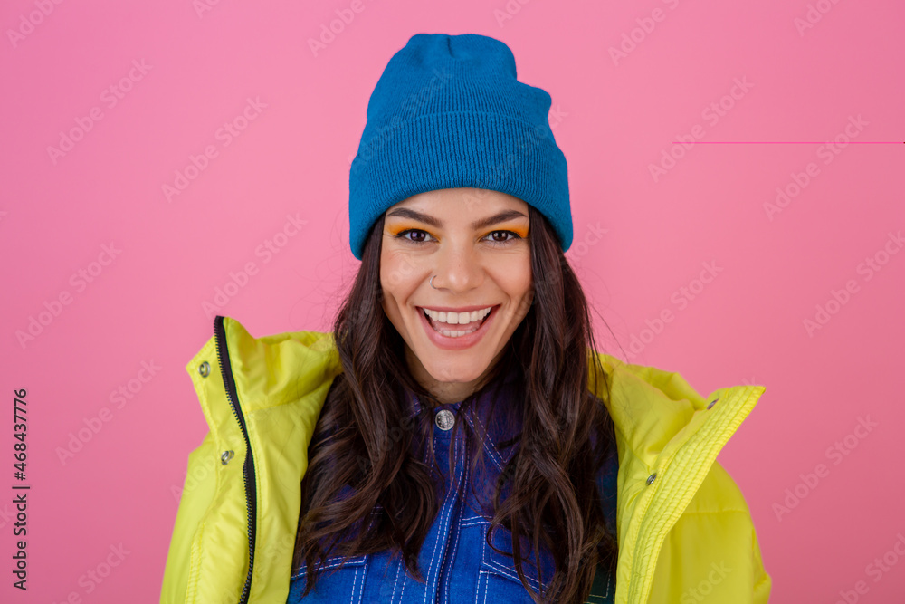 portrait of attractive smiling stylish woman posing in winter fashion look on pink background in bright neon yellow jacket, wearing blue knitted hat, dressed in warm clothes portrait of attractive smiling stylish woman posing in winter fashion look on pink background in bright neon yellow jacket, wearing blue knitted hat, dressed in warm clothes