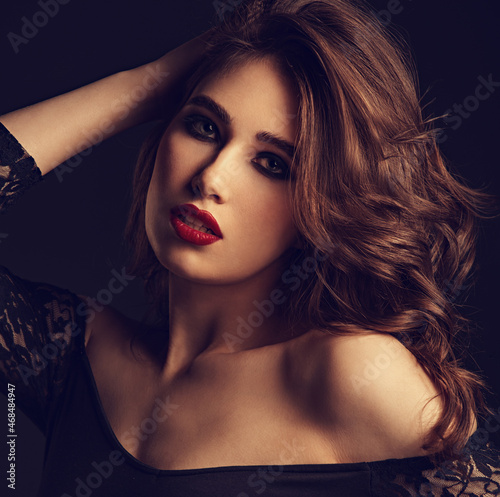 Beautiful bright makeup woman with long brown curly volume hair style, red lipstick, and black smokey eyes looking vamp on black background. Closeup toned color