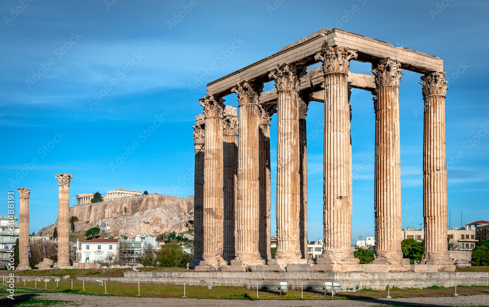 The ruins of the Temple of Olympian Zeus. There are sixteen surviving columns, one of which is lying on the ground. Acropolis is in the background.