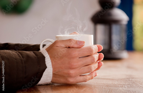 white cup with hot drink and smoke, hands warming up from the heat, at home. Focus on the cup