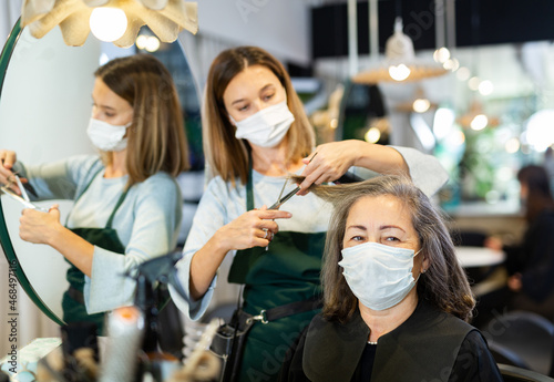 Focused woman hairdresser in protective mask cutting hair of elderly female client in modern hair salon. Pandemic precautionary concept.