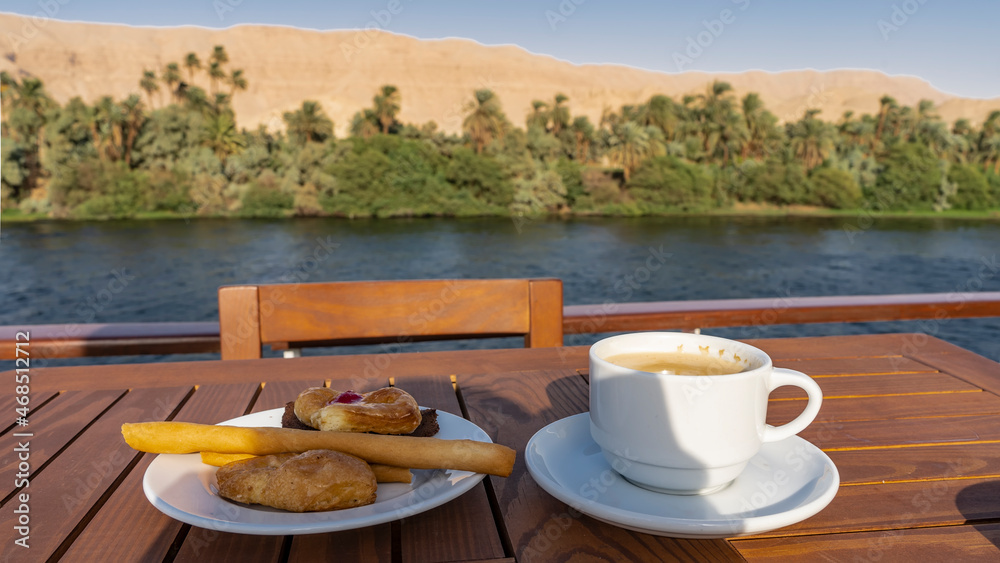 There is a wooden table and chair on the deck of the cruise ship. In a white cup- coffee, cookies on a saucer.  In the background - sand dunes and green vegetation on the banks of the Nile. Egypt
