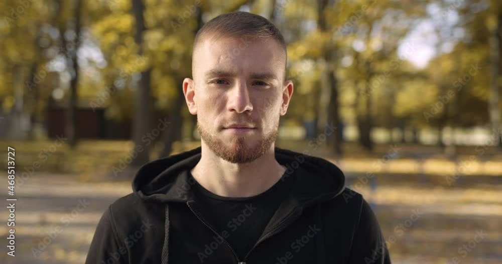 Outdoor portrait of young serious confident caucasian guy wearing hoodie smiling to camera, posing at autumn public park Outdoor portrait of young serious confident caucasian guy wearing hoodie smiling to camera, posing at autumn public park