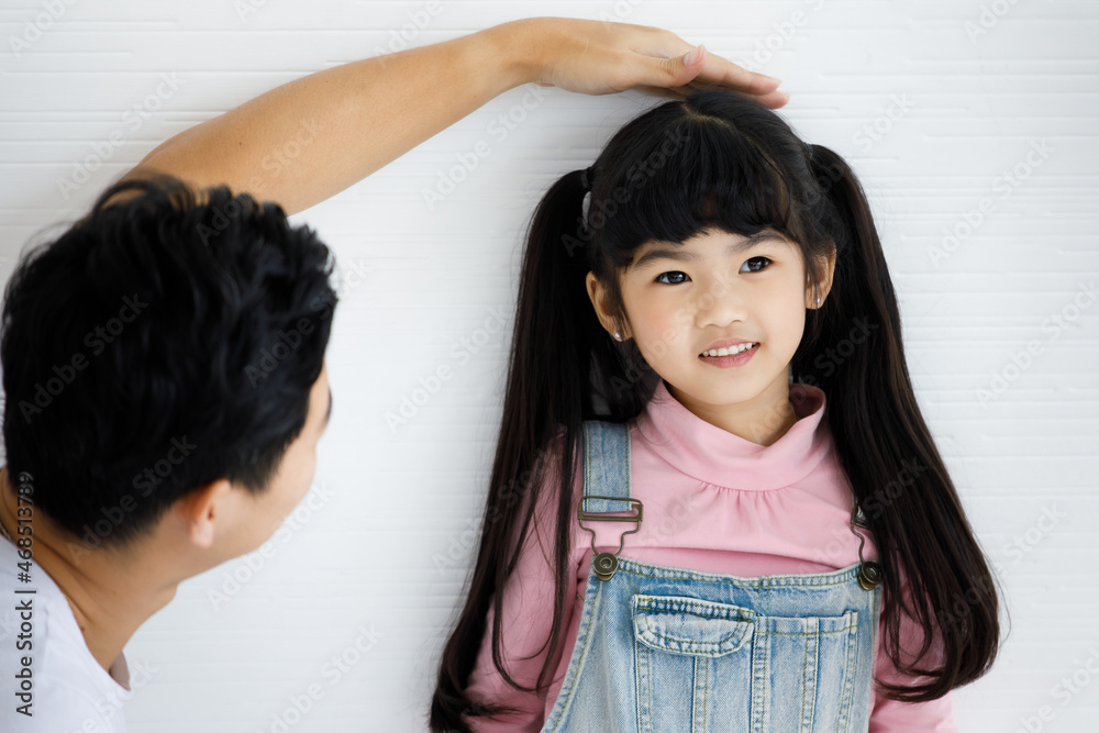 Close up shot of Asian small cute pigtail hair style preschooler child daughter in jeans overalls outfit standing smiling leaning at wall while father using hand measuring checking growth rate height Close up shot of Asian small cute pigtail hair style preschooler child daughter in jeans overalls outfit standing smiling leaning at wall while father using hand measuring checking growth rate height