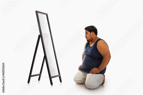 Portrait of a fat man looking at bellyfat mirror in despair against plain background. photo