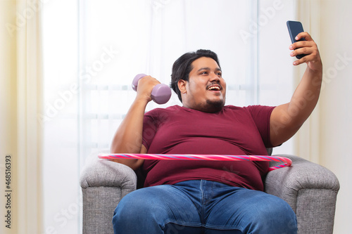 A fat man sitting on couch with dumbles and hulla hoop around his waist clicking selfie. photo