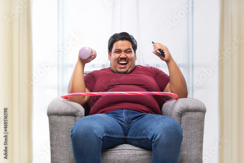 A fat man sitting on couch with dumbles and mobile with a hulla hoop around his waist. photo
