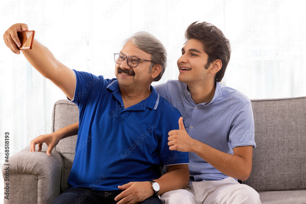 Indian father and son taking selfie while sitting on sofa at home Indian father and son taking selfie while sitting on sofa at home