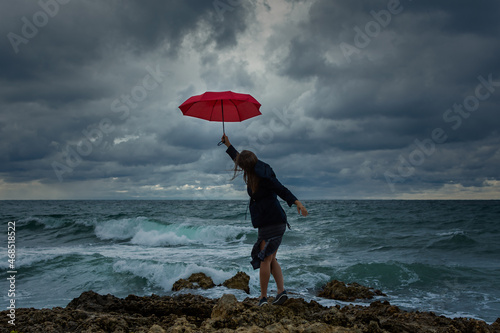 Young woman with a red umbrella on the background of the sea. Atmospheric autumn landscape with a young dark-haired girl in a developing dress. Storm, strong wind, overcast. The concept of happiness