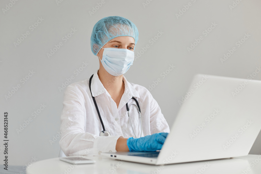 Caucasian woman doctor wearing gown, surgical mask, medical cap and gloves, posing in front of notebook, working working on a new scientific article. Caucasian woman doctor wearing gown, surgical mask, medical cap and gloves, posing in front of notebook, working working on a new scientific article.