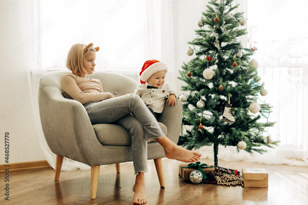 Charming children are sitting in a chair in a room decorated for Christmas and New Year. A boy in a Santa Claus hat and his sister in tiger ears. The concept of Christmas and New Year