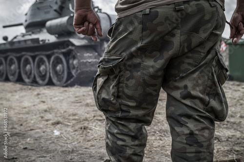 Close-up of soldier's legs on a battlefield, wearing woodland camo military pants (camouflage trousers). With a tank tracked wheels in the background.