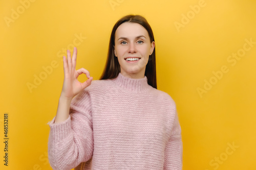 Portrait of smiling pretty happy cute cheerful satisfied positive attractive young woman 20s show ok okay zero finger gesture, wears pink knitted sweater, isolated on yellow color background studio