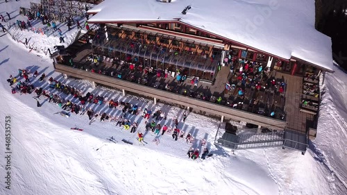 Aerial drone video of crowded ski restaurant with terrace full of people. Apres ski bar surrounded by snow with stage and live music cramped with skiers relaxing. Mountain hut overcrowded by tourists. photo