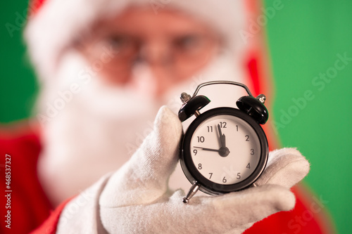 Santa Claus in red coat and hat against dark green background, wearing white gloves and showing a small black alarm clock in his hand; two minutes till midnight