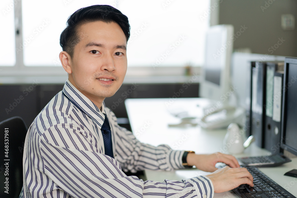 Portrait of cheerful Asian male employee working on computer at desk in the office looking at camera. worker life in workplace. copy space Portrait of cheerful Asian male employee working on computer at desk in the office looking at camera. worker life in workplace. copy space