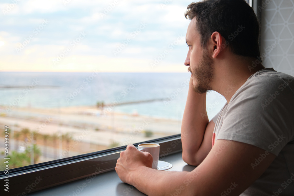 Young caucasian man drink coffee look at the sunrise and sea throw the window.