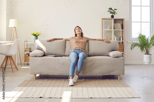 Happy relaxed young woman resting sitting on comfortable sofa in living room at home. Portrait of girl in casual clothes enjoying lazy weekend or vacation relaxing in cozy room of her home. photo